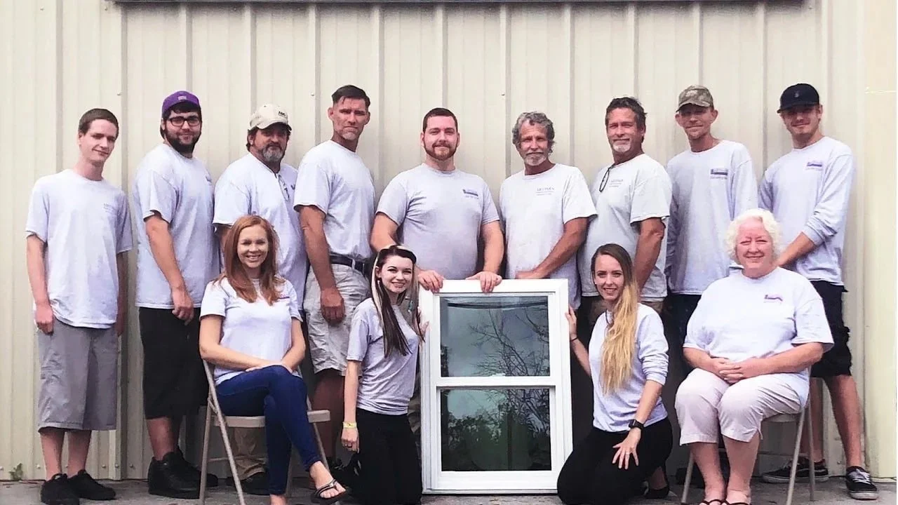 The Artisan Windows & Doors crew — thirteen team members in Artisan shirts, gathered around a replacement window.